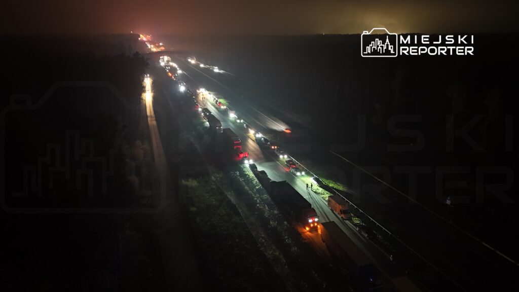 Drogą krajową poruszają się pojazdy, tworząc długi korek w nocy, otoczone przez lasy.