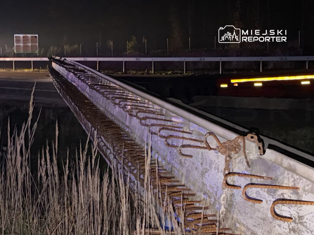 Metalowy element konstrukcji leżący na poboczu drogi, otoczony wysoką trawą, w nocy oświetlony przez latarnie.