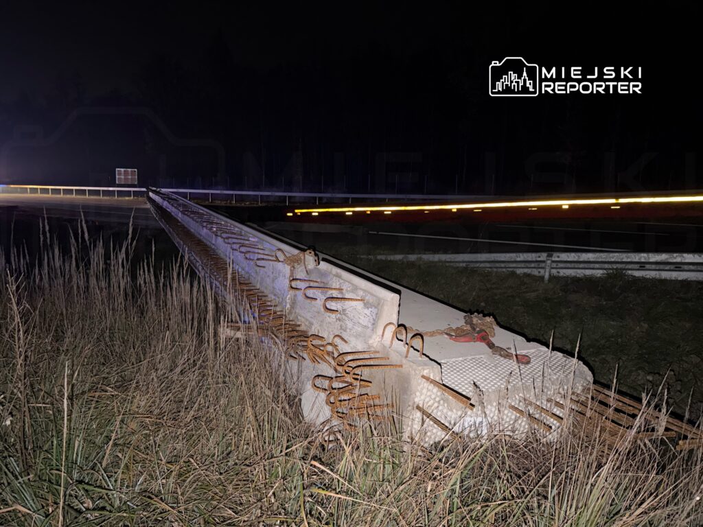 Zardzewiały element betonowy leży na poboczu drogi, otoczony wysoką trawą, w nocy z widocznymi światłami samochodów w tle.