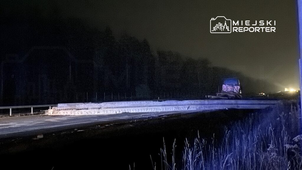 W nocy ciężki sprzęt budowlany pracuje na odcinku drogi, otoczony przez ciemność i zarośla.