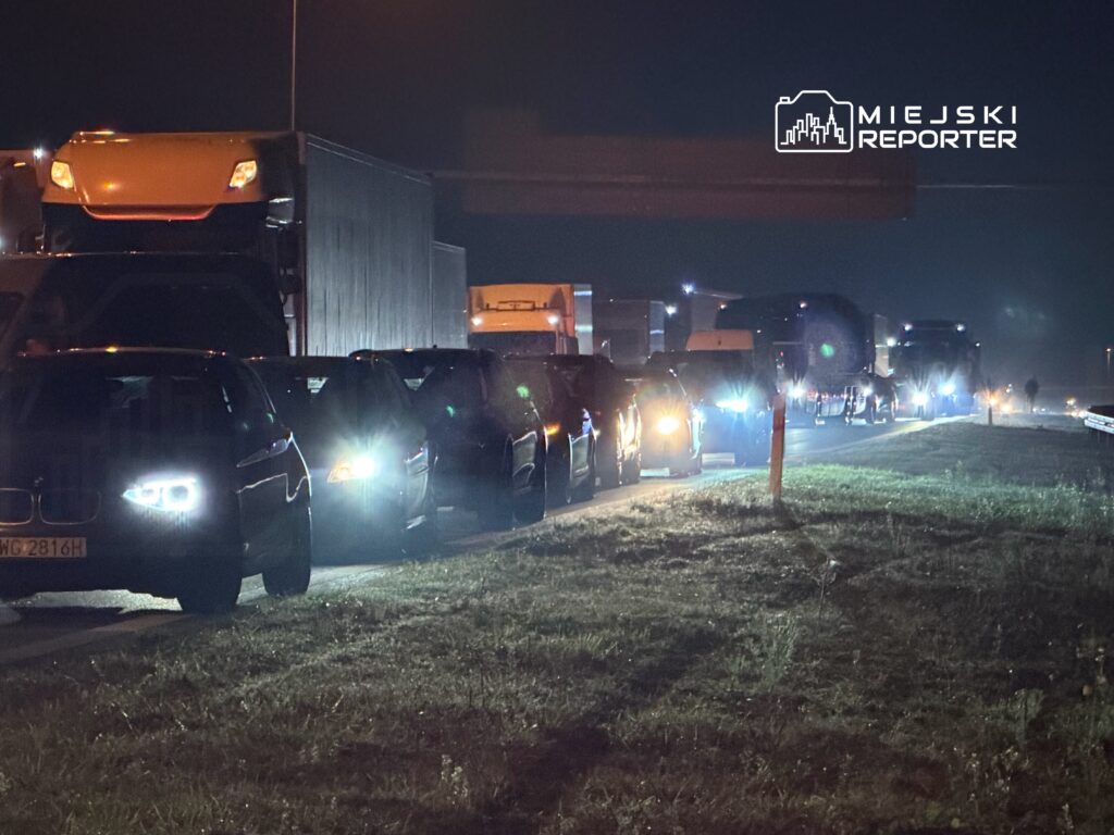 Kolejka samochodów osobowych i ciężarowych stojących na drodze w nocy, oświetlonych przez reflektory.