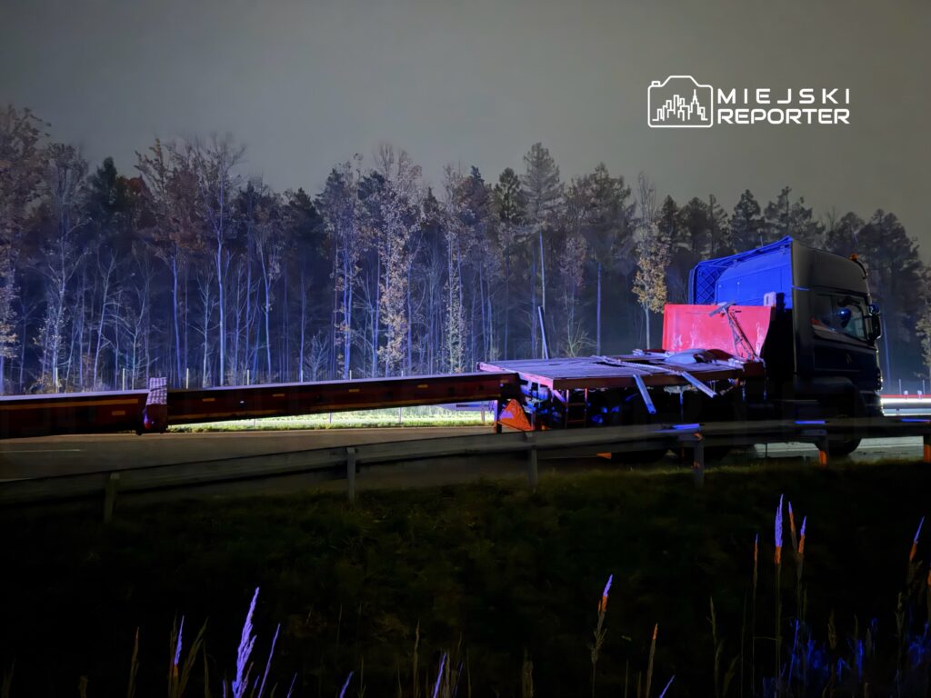 Ciężarówka z uszkodzonym ładunkiem stoi na poboczu drogi, w tle widoczne drzewa oświetlone niebieskim światłem.