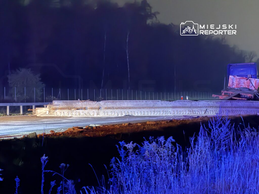 Droga budowlana z betonowymi elementami w tle, oświetlona niebieskim światłem, w otoczeniu niskiej roślinności.