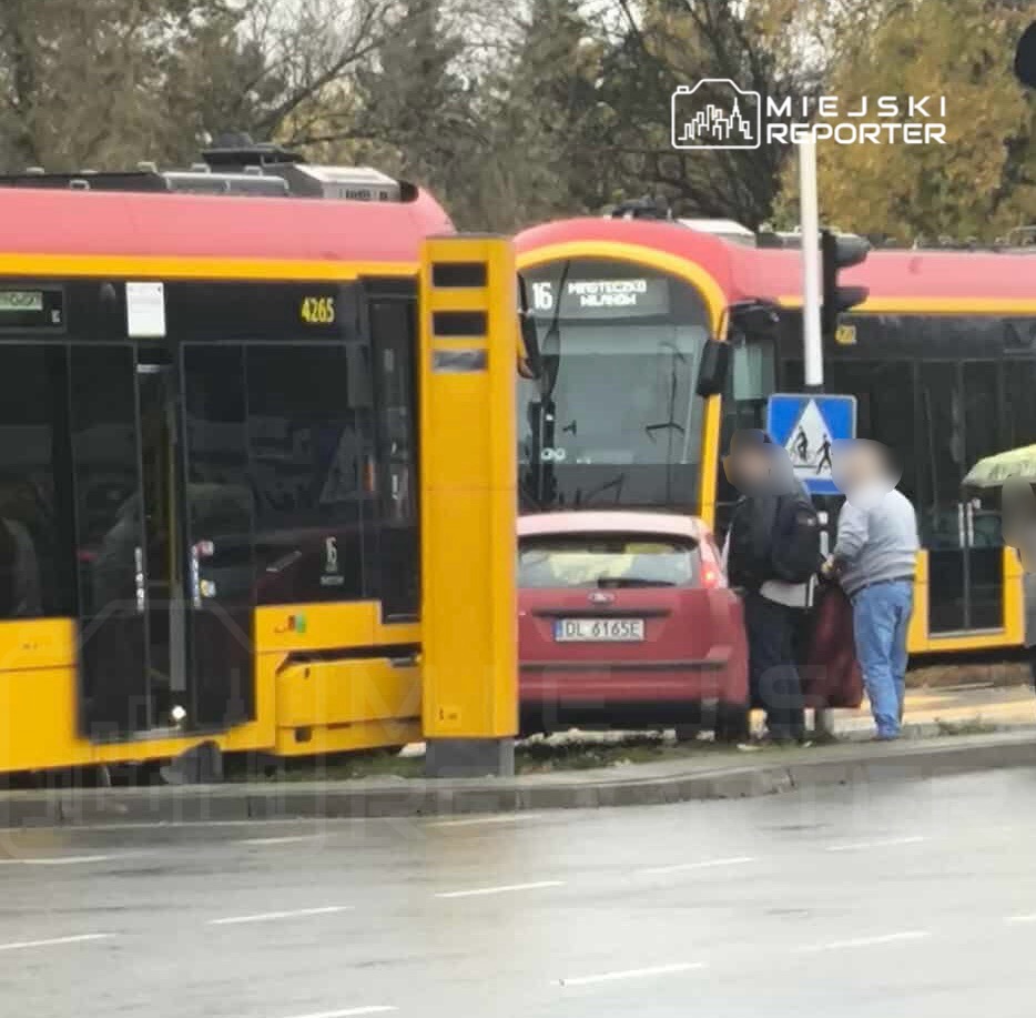 Czerwony samochód osobowy stoi w zatorze między dwoma autobusami, a dwie osoby rozmawiają obok pojazdu.