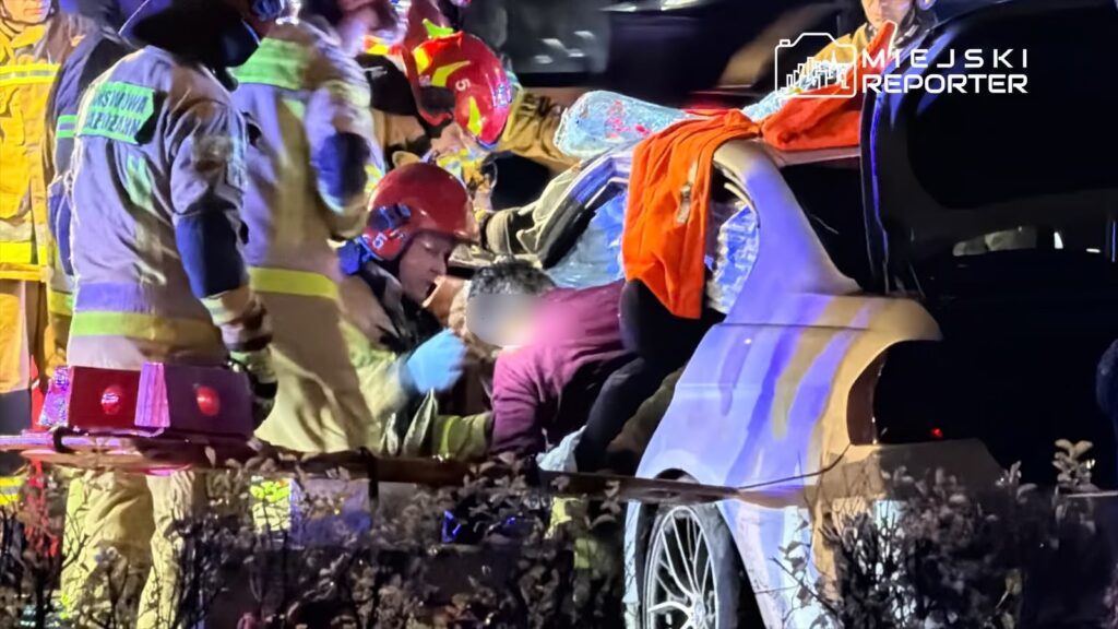Ratownicy medyczni i strażacy pomagają osobie wydobytej z wraku samochodu w nocy, otoczeni przez sprzęt ratunkowy.
