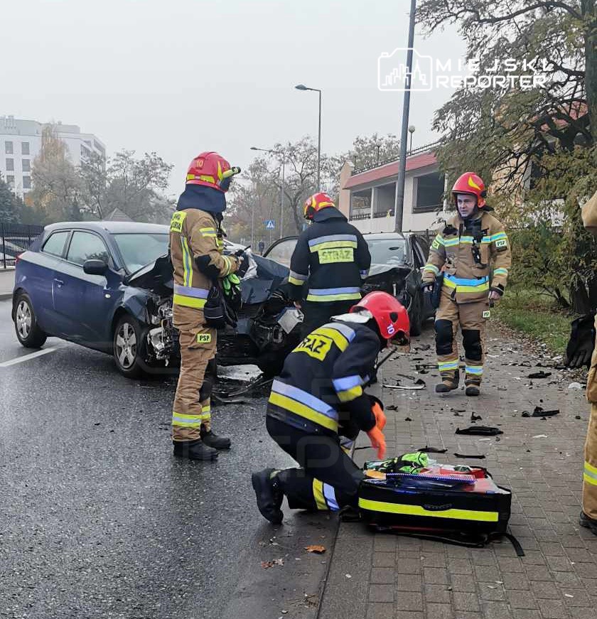 Czterech strażaków pracuje nad wypadkiem drogowym, jeden z nich przygotowuje sprzęt medyczny obok uszkodzonego samochodu.