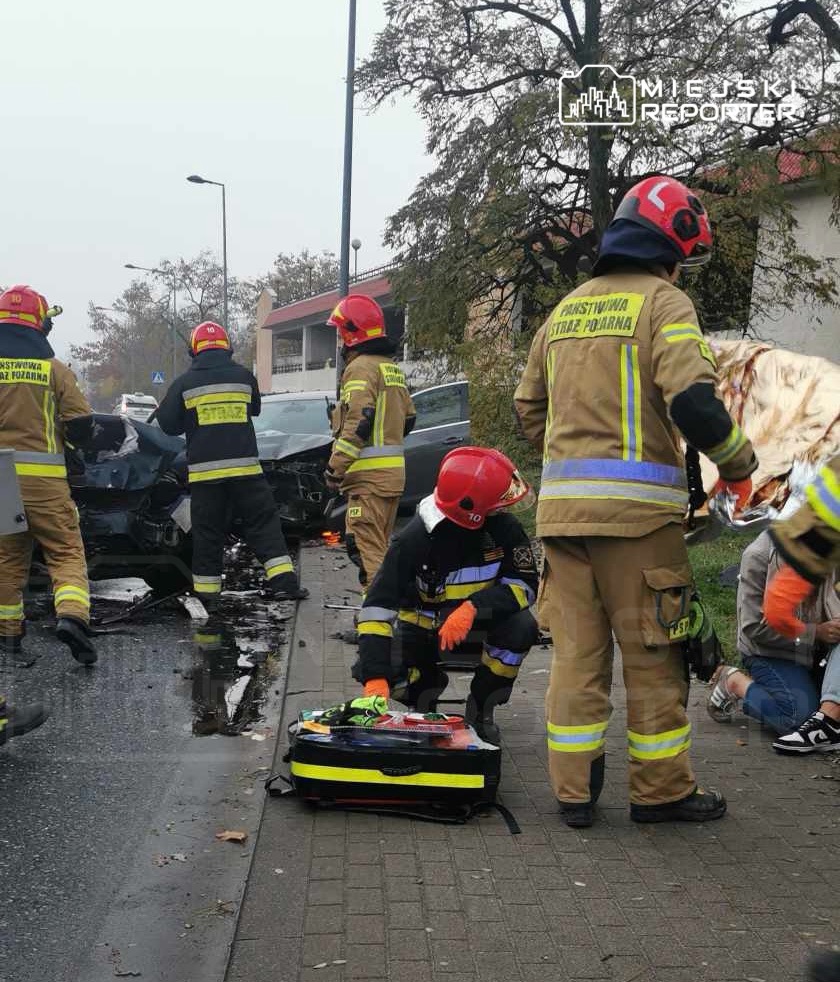 Grupa strażaków w hełmach i mundurach pracuje przy wypadku drogowym, a jeden z nich przygotowuje sprzęt ratunkowy.