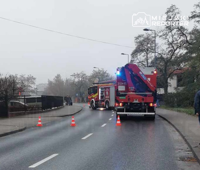 Straż pożarna z włączonymi sygnałami świetlnymi blokuje drogę, a obok stoją ludzie w deszczowej aurze.