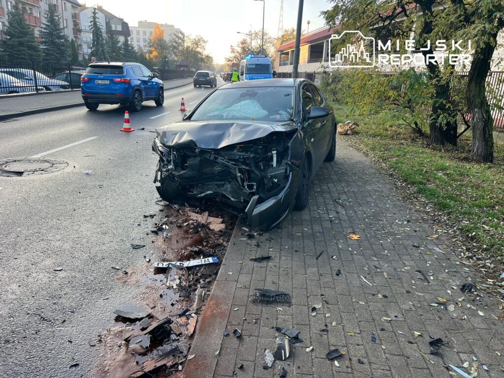 Czarny samochód osobowy z uszkodzoną przednią częścią stoi na poboczu, wokół leżą fragmenty karoserii.