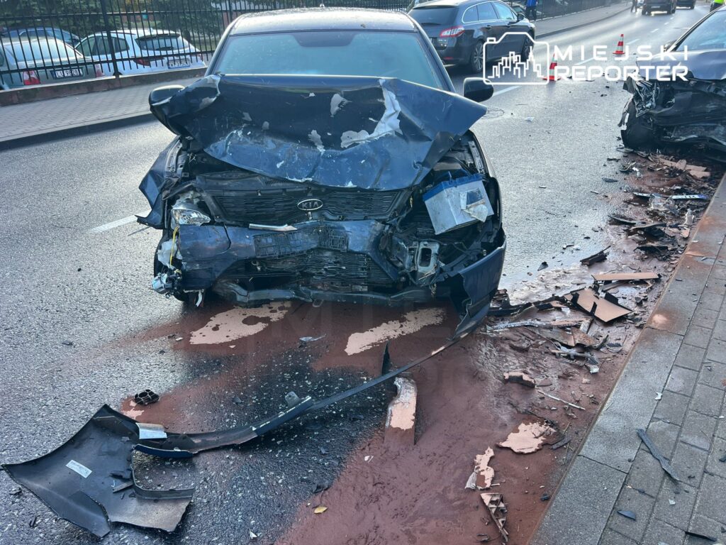 Zniszczony przód samochodu osobowego na drodze, z częściami leżącymi obok i rozlanym płynem na jezdni.