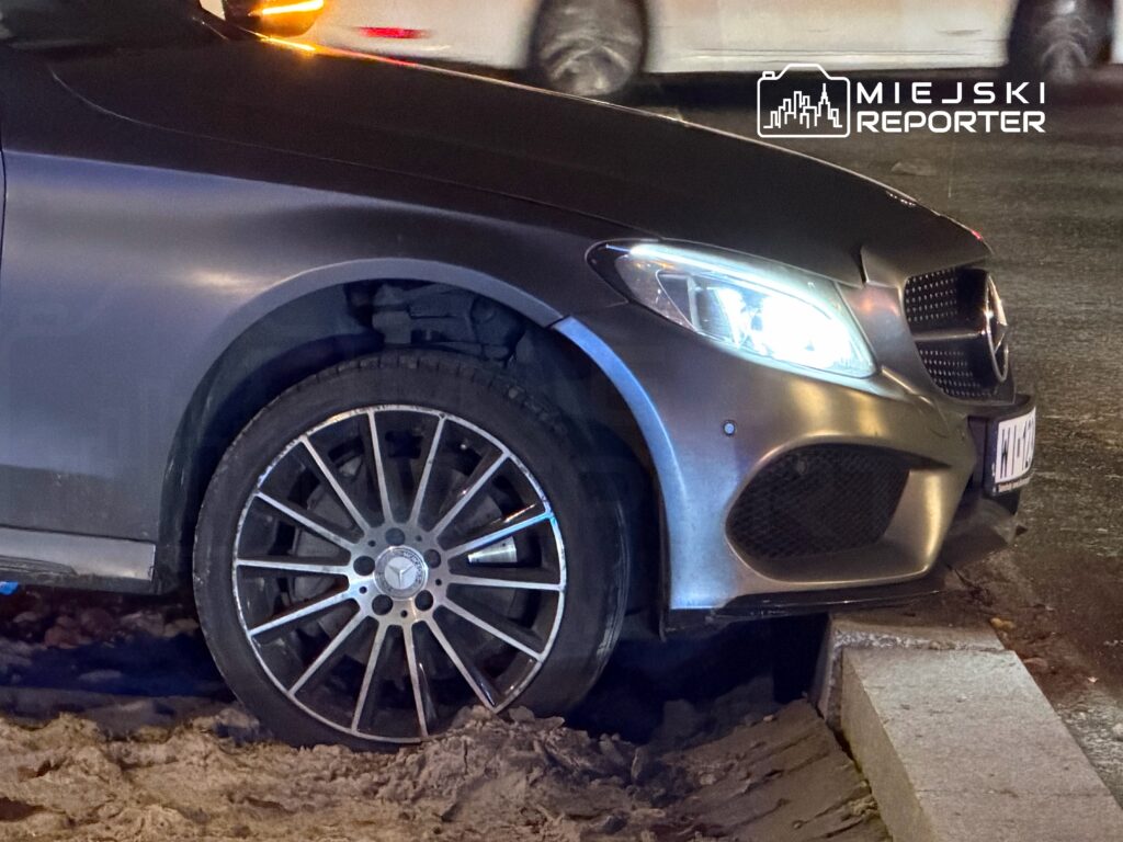 Samochód osobowy marki Mercedes z uszkodzoną oponą zaparkowany na krawężniku w zaśnieżonym otoczeniu.