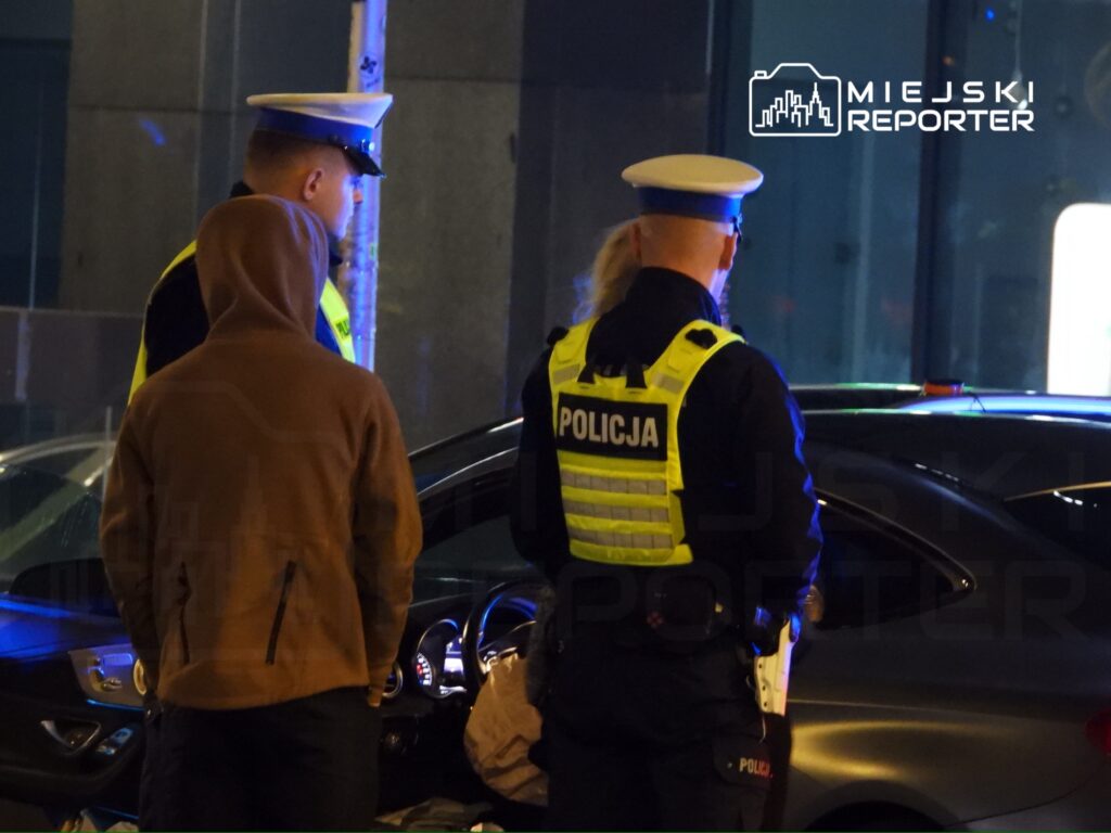 Dwóch policjantów w odblaskowych kamizelkach rozmawia z osobą w kapturze przy zaparkowanym samochodzie.