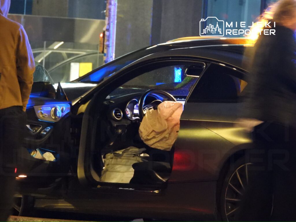 Samochód z otwartymi drzwiami, w środku widać wystrzeloną poduszkę powietrzną, obok przechodnie w ciemnej scenerii.