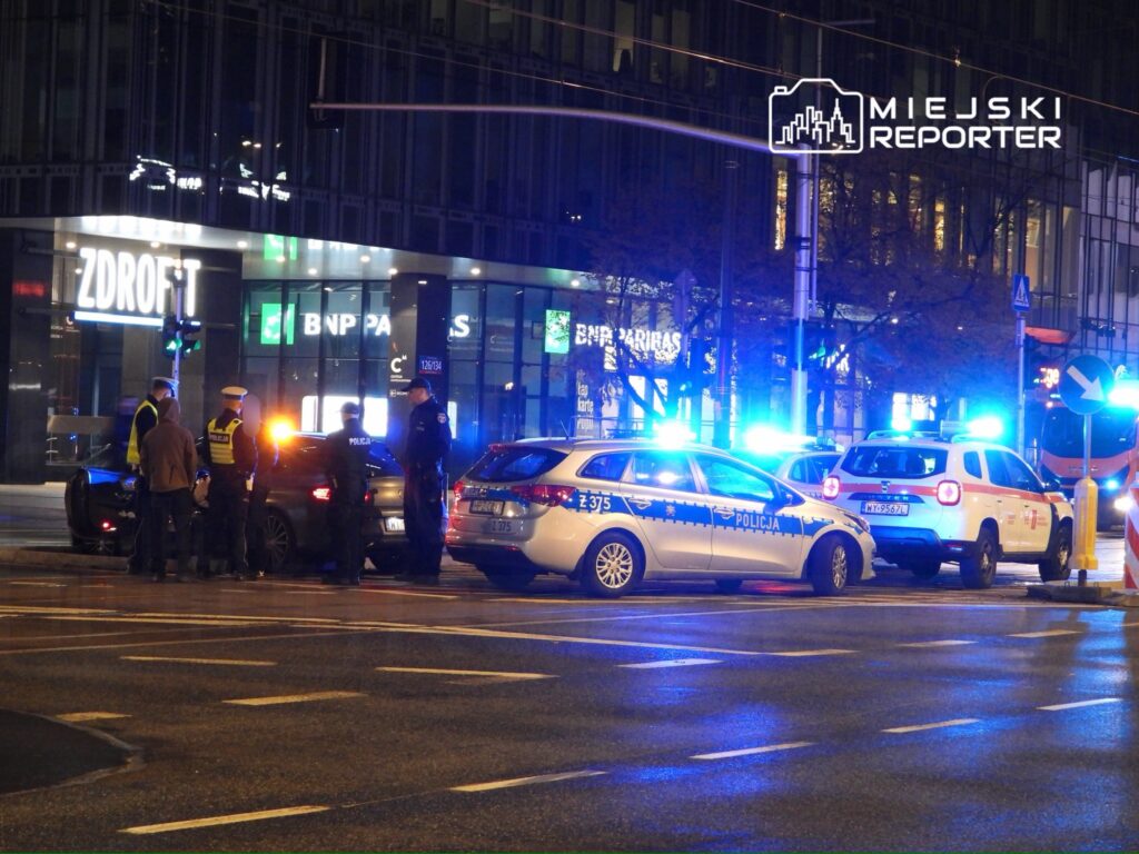 Grupa policjantów rozmawia z kierowcą przy zatrzymanym samochodzie na ulicy, w tle widać radiowozy z włączonymi sygnałami świetlnymi.