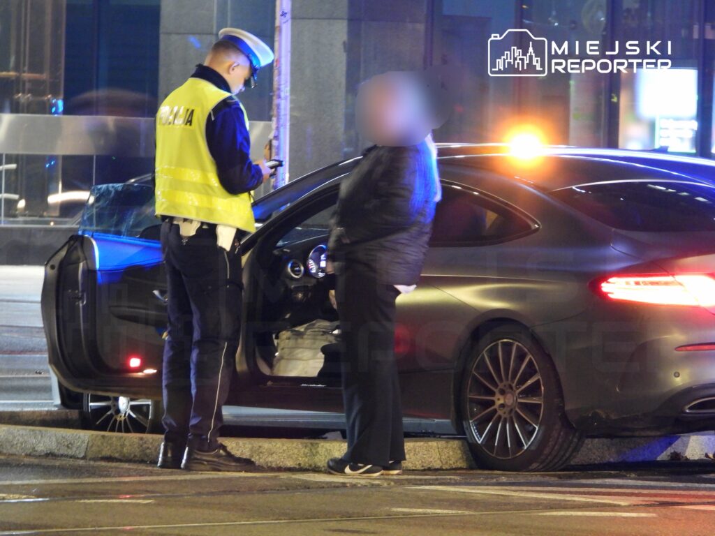 Policjant w żółtej kamizelce sprawdza dokumenty kierowcy przy otwartym drzwiach czarnego samochodu osobowego.