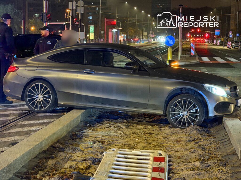 Samochód osobowy stoi na torach tramwajowych, podczas gdy policjanci rozmawiają w tle. Nocna sceneria z oświetlonymi ulicami.