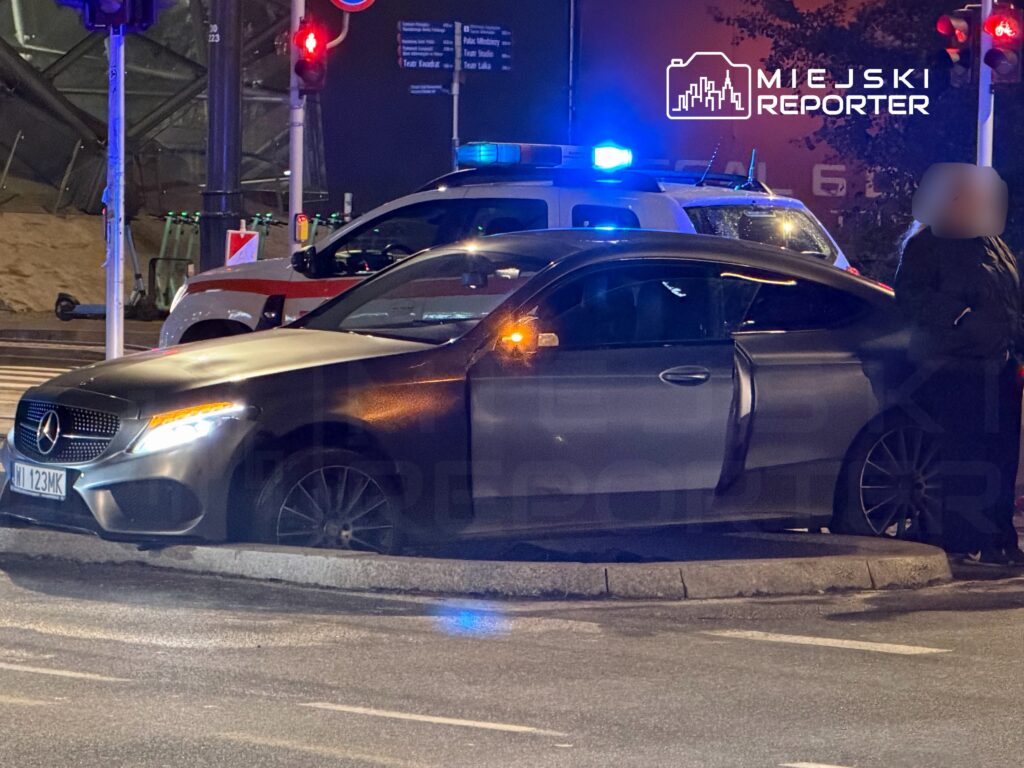 Czarny samochód marki Mercedes zatrzymany przy sygnalizacji świetlnej, obok radiowozu policyjnego z włączonymi światłami.