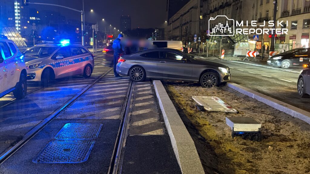 Policyjny radiowóz z niebieskimi światłami stoi obok samochodu osobowego na torowisku w nocy, w tle widać ruch uliczny.