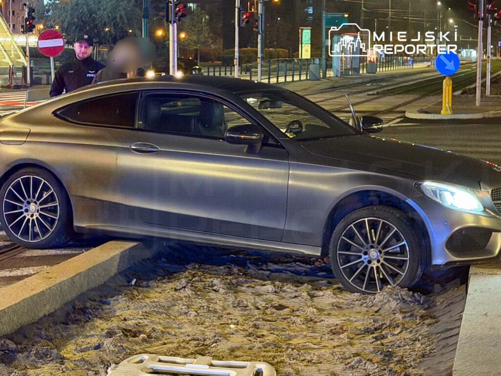 Mężczyzna w mundurze policjanta stoi obok samochodu osobowego na krawężniku, w tle oświetlona ulica.