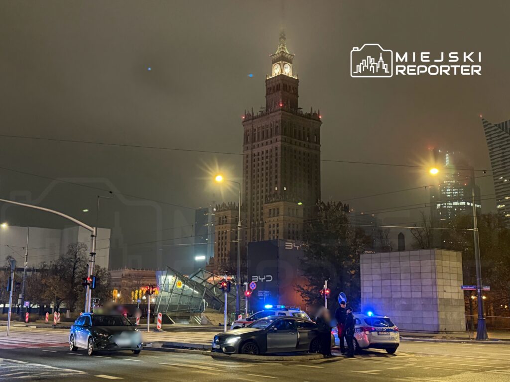 Dwa samochody osobowe zatrzymane na skrzyżowaniu, obok policyjne radiowozy z włączonymi sygnałami świetlnymi, w tle wieżowiec.