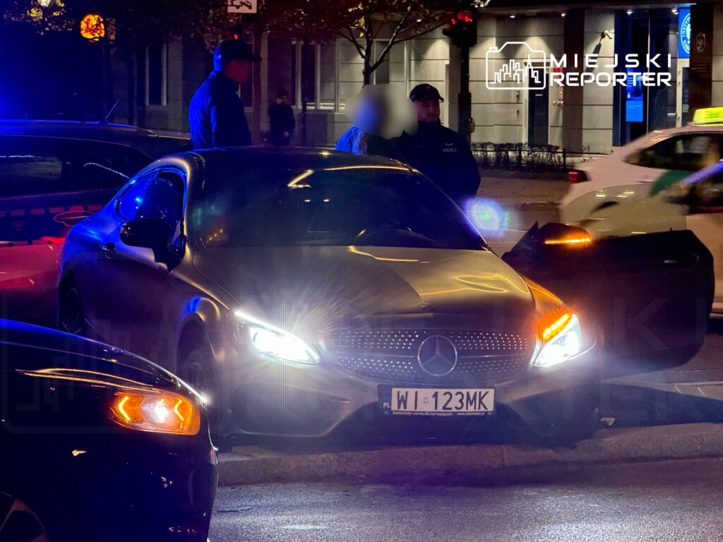 Mężczyzna w mundurze policjanta stoi obok zatrzymanego samochodu osobowego marki Mercedes na oświetlonej ulicy.