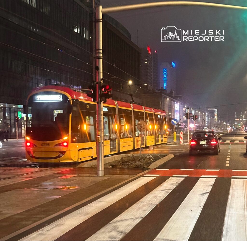 Tramwaj w żółto-czerwonym kolorze przejeżdża przez skrzyżowanie w oświetlonym mieście nocą.