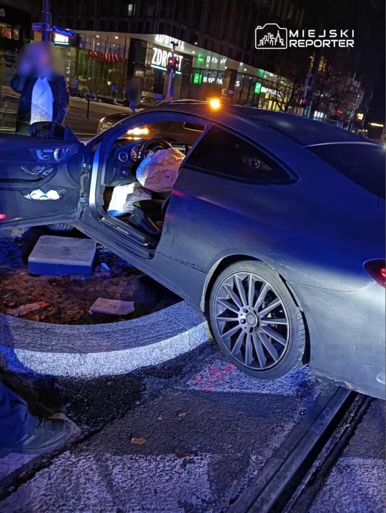 Samochód osobowy utknął w głębokim wykopie drogowym, a w jego wnętrzu widoczne są poduszki powietrzne. W tle widać oświetlenie miejskie.