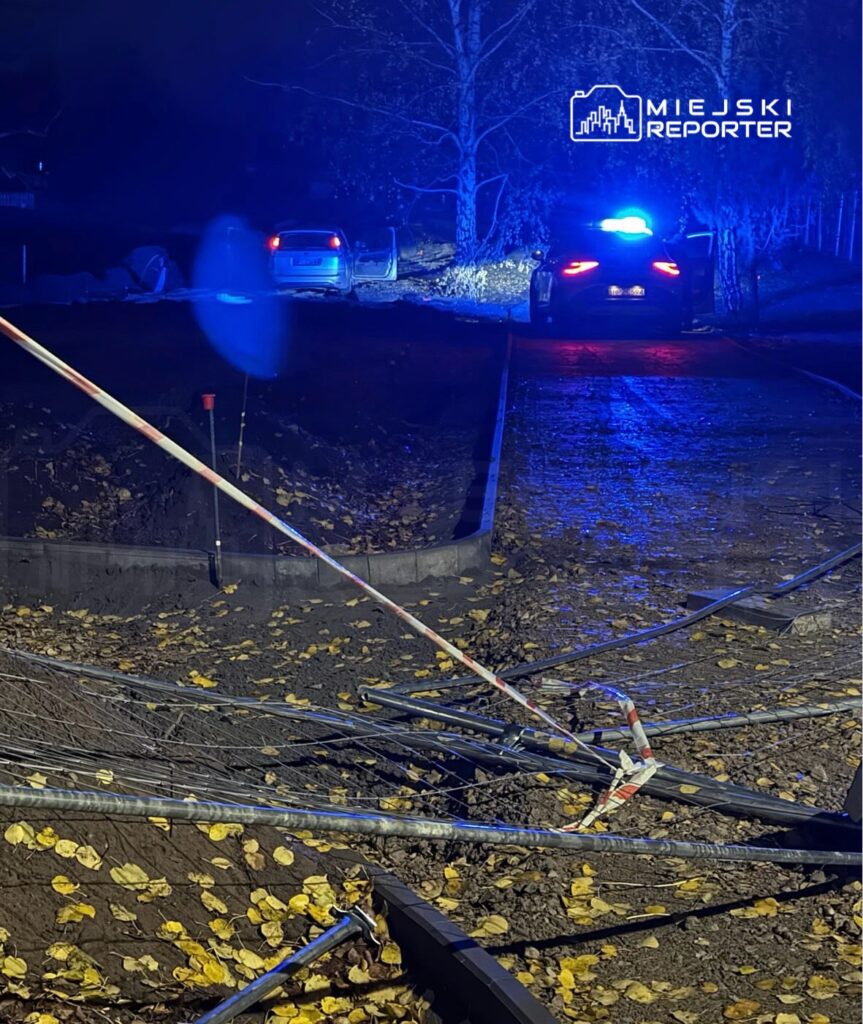 Zablokowana droga z powalonymi słupami i leżącymi gałęziami, w tle radiowóz policyjny z włączonymi sygnałami świetlnymi.