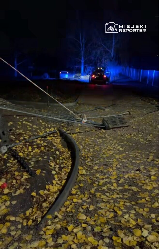Uszkodzona linia tramwajowa leży na drodze pokrytej żółtymi liśćmi, a w tle widać niebieskie światła radiowozu.