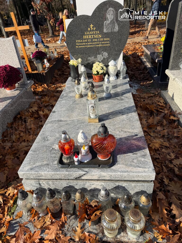 Grób z ciemnoszarym nagrobkiem w kształcie serca, ozdobiony zniczami i kwiatami, na tle opadłych liści.