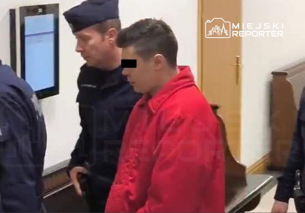 Mężczyzna w czerwonej kurtce prowadzony przez policjanta w budynku sądu.