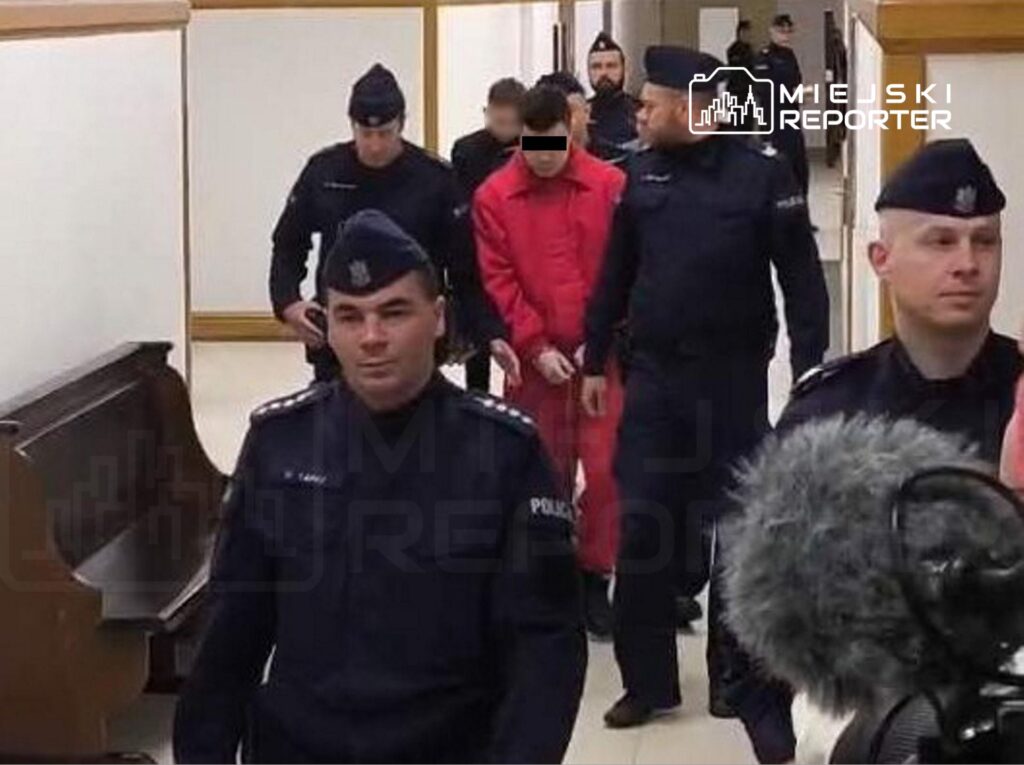 Mężczyzna w czerwonym uniformie eskortowany przez policjantów w korytarzu budynku sądowego.