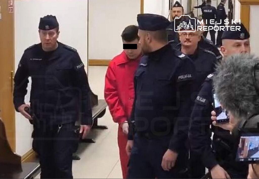 Mężczyzna w czerwonym kombinezonie prowadzony przez policjantów w korytarzu sądowym.