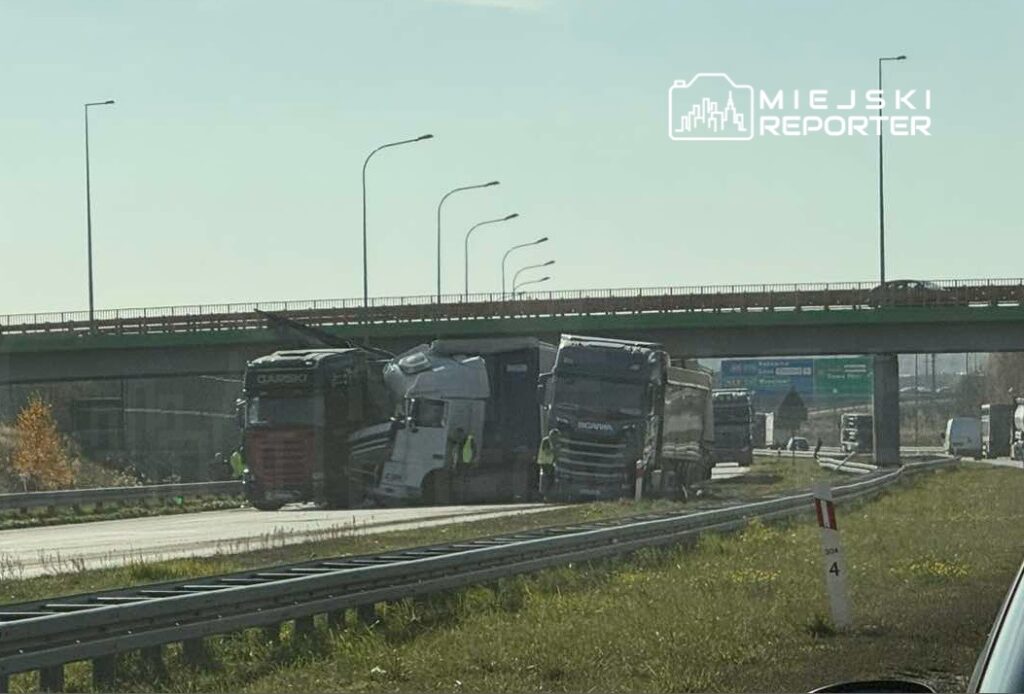 Zestaw ciężarówek zderzonych na drodze pod wiaduktem, z widocznymi uszkodzeniami i interwencją służb.