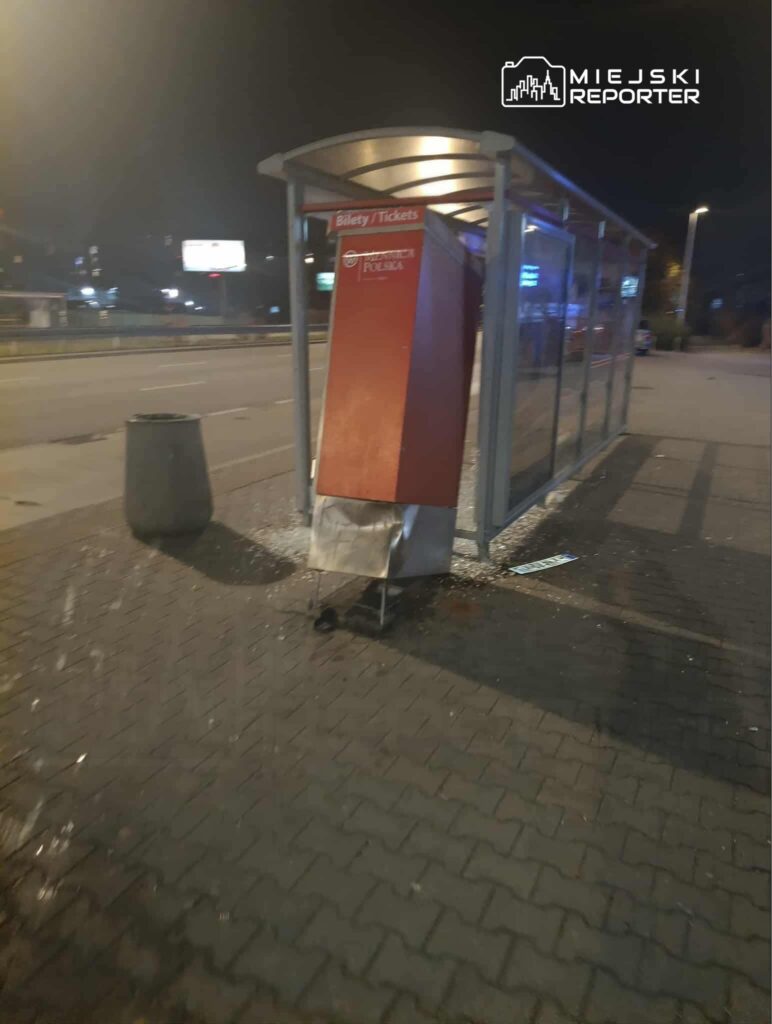 Uszkodzony automat biletowy umieszczony w zadaszonej wiacie przystankowej na nocnej ulicy.