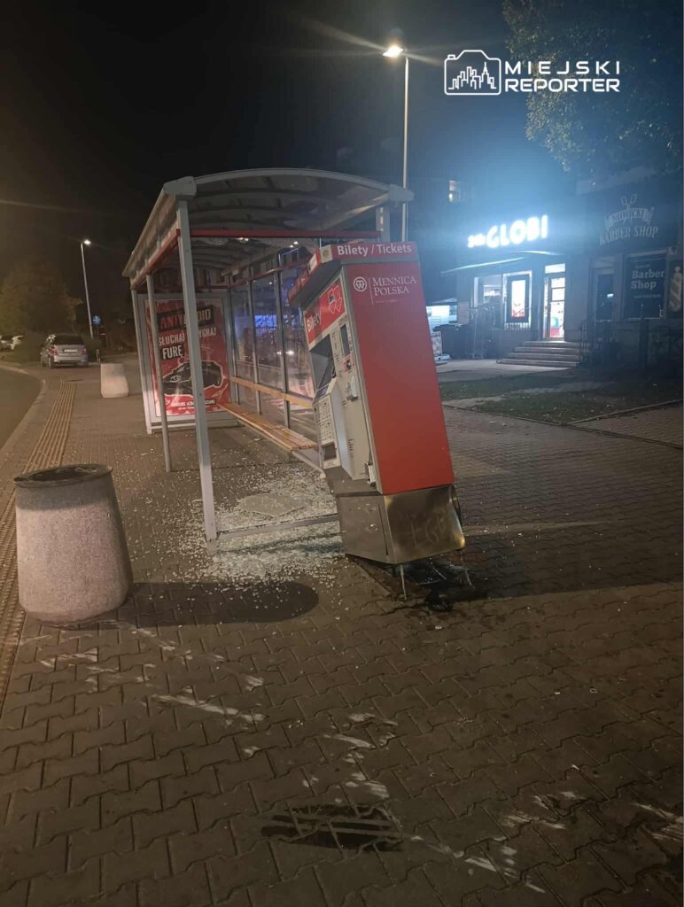 Uszkodzony automat biletowy stoi przy przystanku, obok rozbite szkło, w tle widoczny sklep nocny.