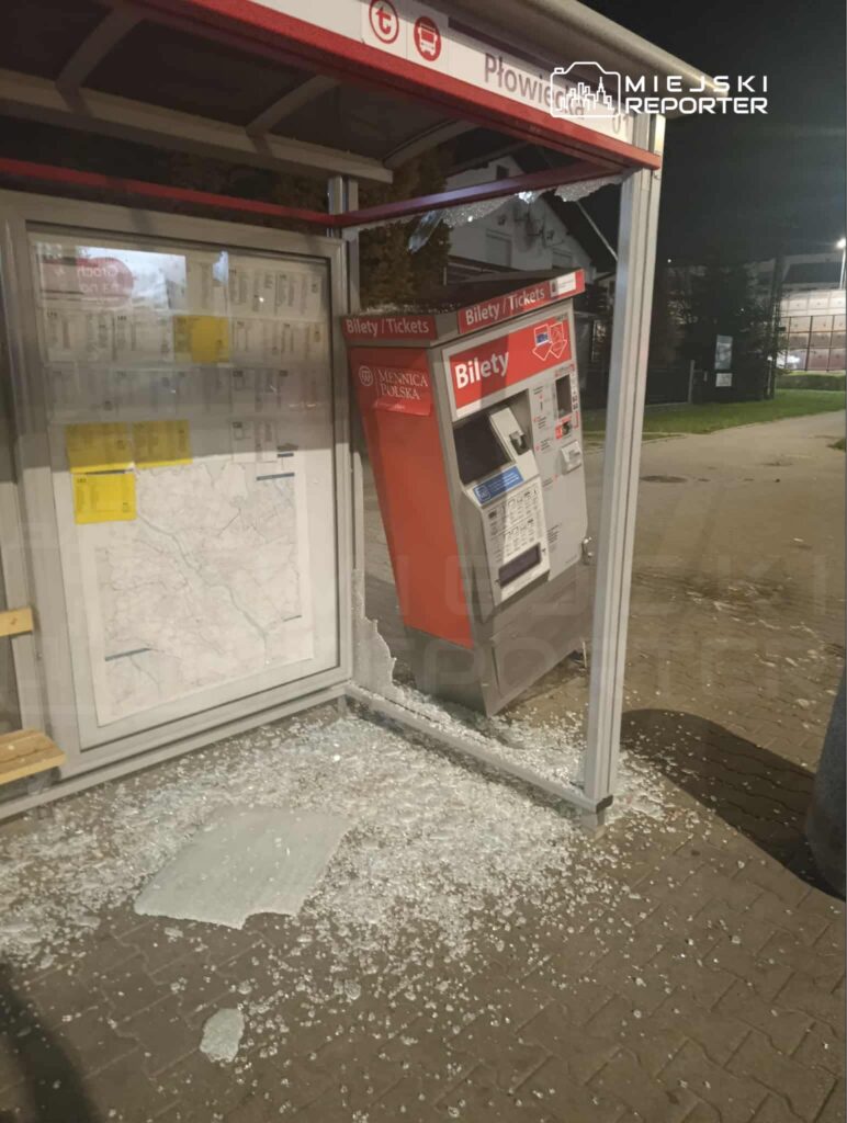 Zniszczony automat biletowy przy przystanku, z rozbitym szkłem leżącym na chodniku. W tle widoczna mapa komunikacyjna.