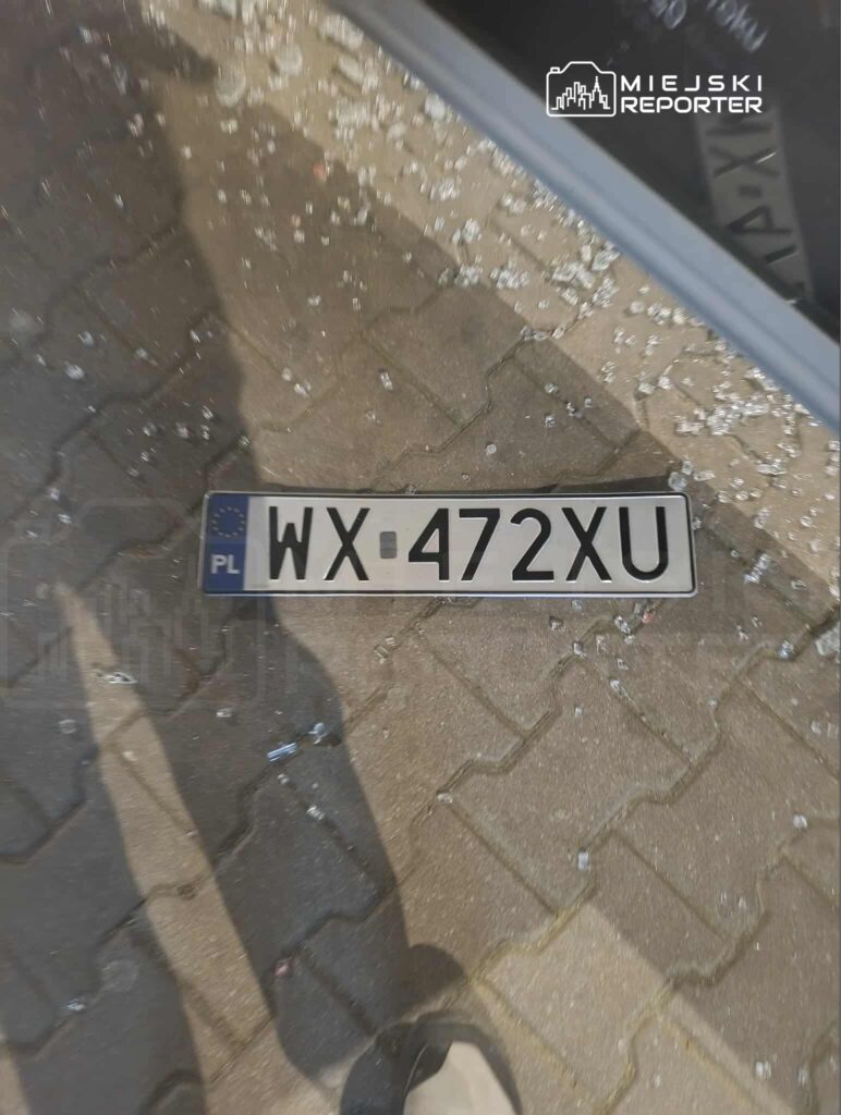 Na chodniku leży uszkodzona tablica rejestracyjna z numerem WX-472XU, otoczona kawałkami szkła.