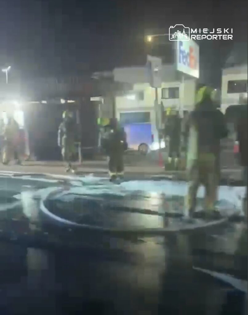 Grupa strażaków w hełmach pracuje nocą na miejscu akcji, oblewając podłoże pianą gaśniczą.