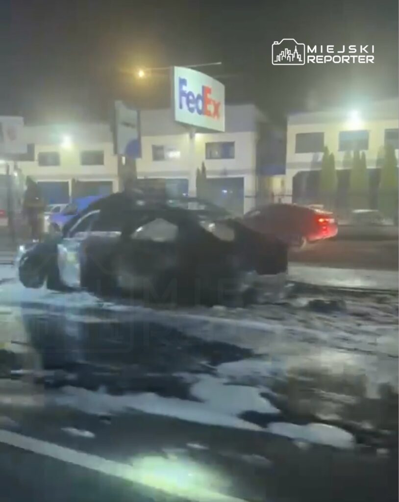 Samochód leży na boku na drodze, obok widać ślady wody i piany, w tle znak FedEx oraz inne pojazdy.