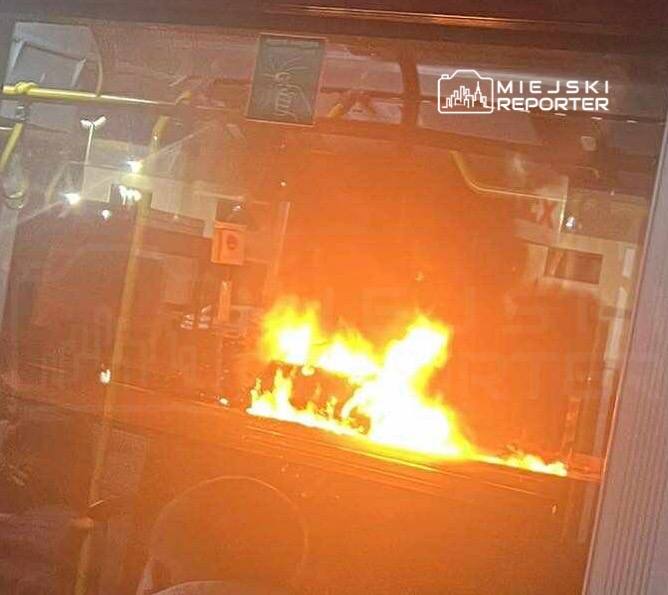 Płonący pojazd komunikacji miejskiej widoczny przez okno, z intensywnym ogniem wydobywającym się z wnętrza.