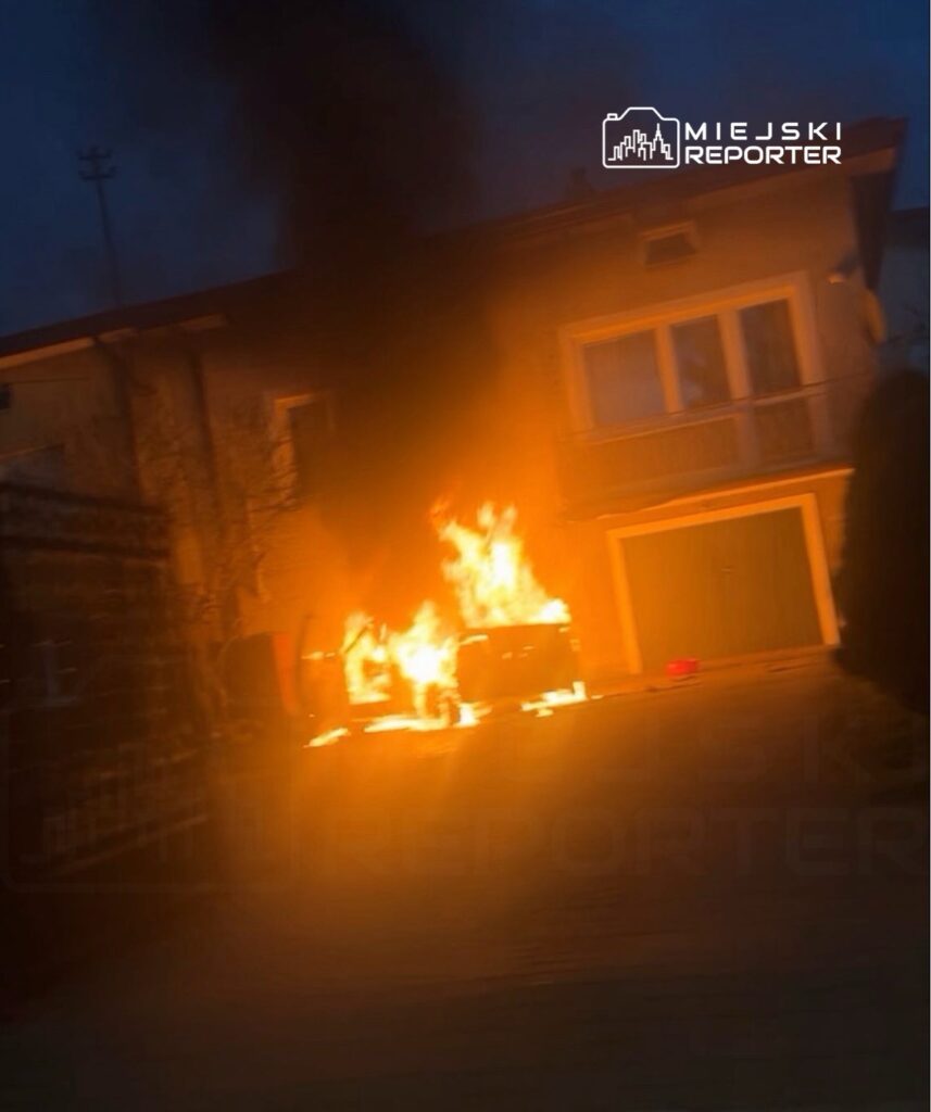 Pojazd zaparkowany przed budynkiem płonie, wydobywa się z niego czarny dym w ciemnej scenerii nocnej.