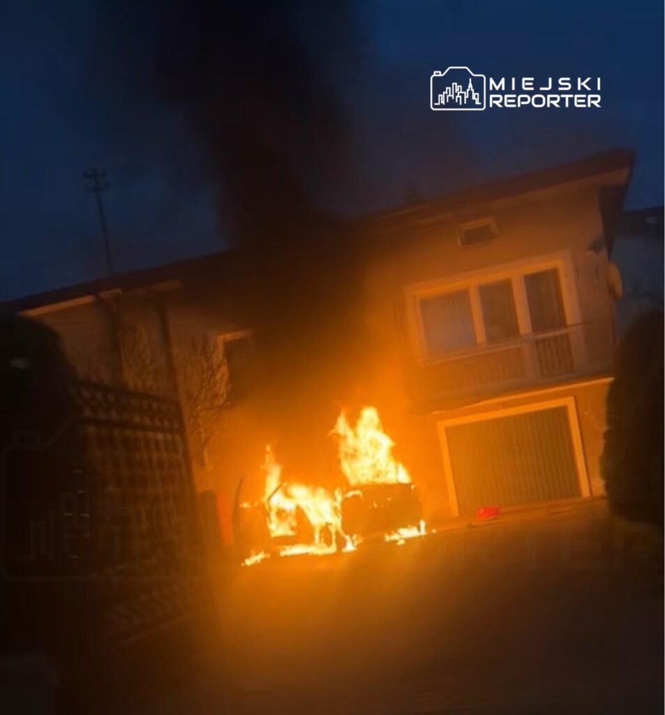 Samochód płonący na podjeździe przed budynkiem mieszkalnym, wydobywający się czarny dym.