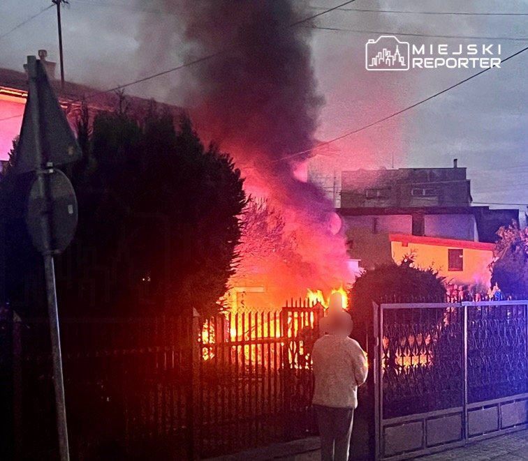 Kobieta obserwująca pożar budynku, z którego wydobywa się czarny dym, w otoczeniu ogrodzenia i drzew.