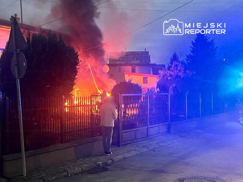 Mężczyzna w jasnej odzieży obserwujący pożar w pobliżu ogrodzenia, w tle widoczne płomienie i dym.