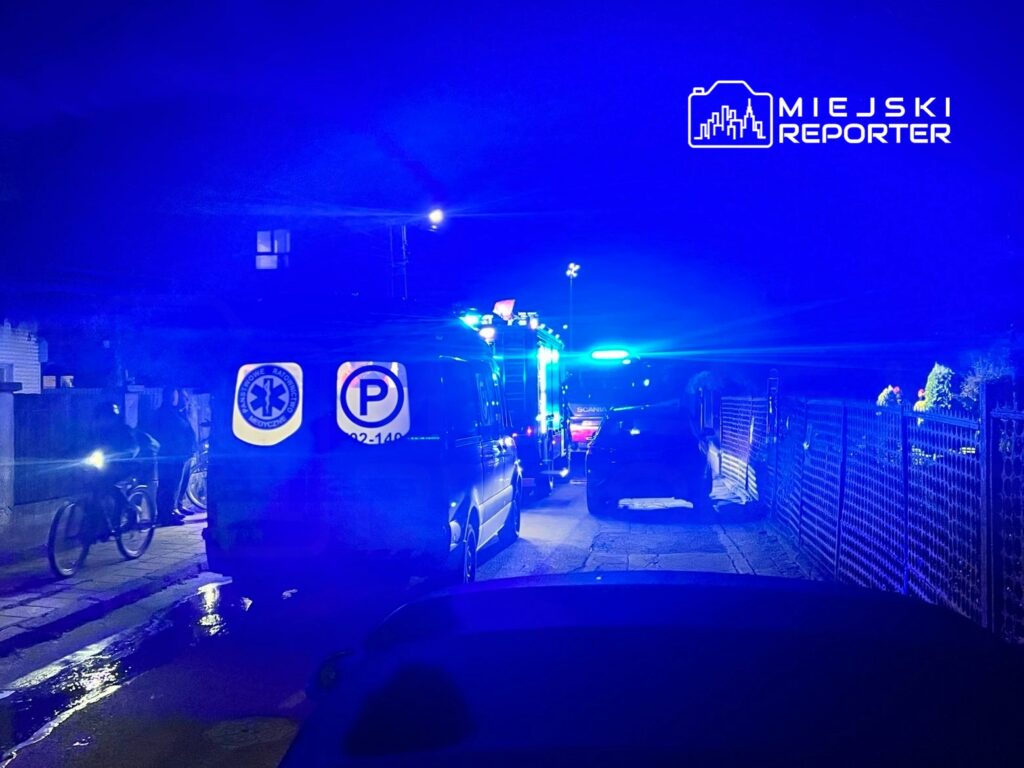 Zespół ratunkowy z niebieskimi światłami zatrzymany na wąskiej ulicy, obok rowerzysty.