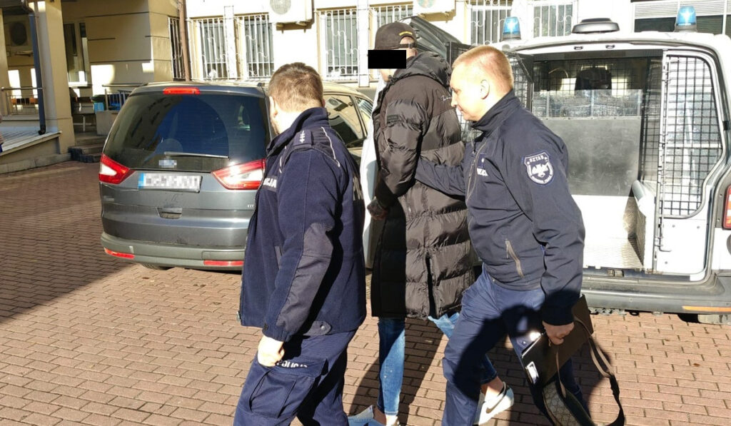 Mężczyzna w długim płaszczu prowadzony przez dwóch policjantów na zewnątrz budynku, obok zaparkowanego samochodu.