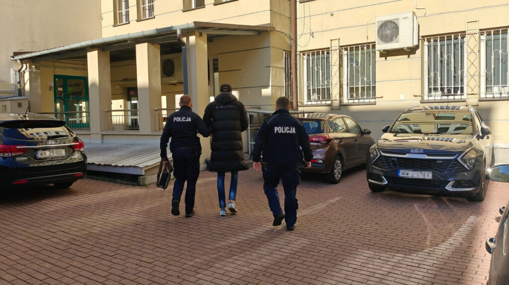 Dwaj policjanci prowadzą mężczyznę w czarnej kurtce przez parking przed budynkiem.