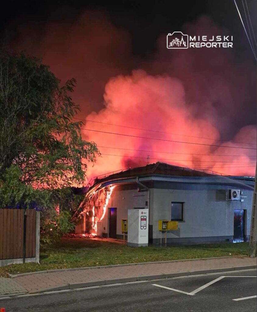 Płonący budynek z widocznymi płomieniami i dymem, otoczony drzewami i ulicą.