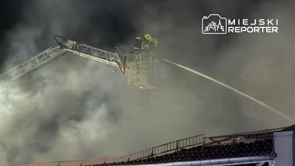 Strażak w podnośniku gaśniczym polewa wodą miejsce pożaru, otoczone gęstym dymem.